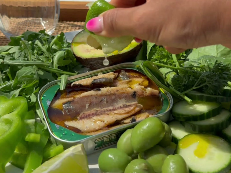 An open tin of sardines surrounded by avocado, rocket, cucumber, green olives and lime